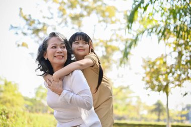 Happy and charming Asian grandmother and her lovely granddaughter in the park, piggy back, spending time together, happy family time. family bonding concept