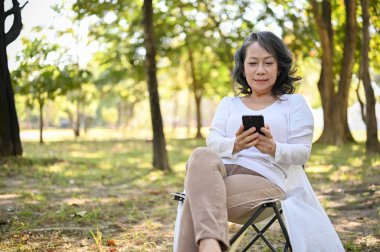 Charming and happy Asian 60s aged woman in casual clothes using her smartphone, chatting with someone while relaxing in the park.