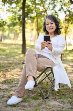 Portrait, Beautiful and charming Asian aged woman in casual clothes using her smartphone, relaxing in the park.