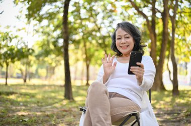 Happy and cheerful Asian 60s aged woman in comfy clothes talking on video call with her grandchild, waving hand to say hi, relaxing in the park.