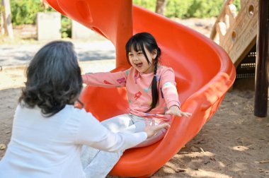 Happy and overjoyed Asian little girl or granddaughter playing on slider, enjoys playing on playground with her grandmother in the park. healthy and fun outdoor activity for kids concept