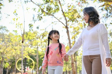 Happy and lovely Asian granddaughter holding hand with her grandmother while strolling in the beautiful park together. happy family time concept