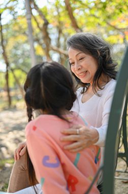 Caring and kind Asian grandmother tells a story to her granddaughter while relax on a bench in the park together.