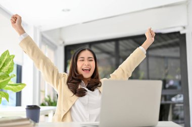 Carefree and overjoyed millennial Asian businesswoman or female office worker raising hands, screaming with happiness, celebrating her career success.