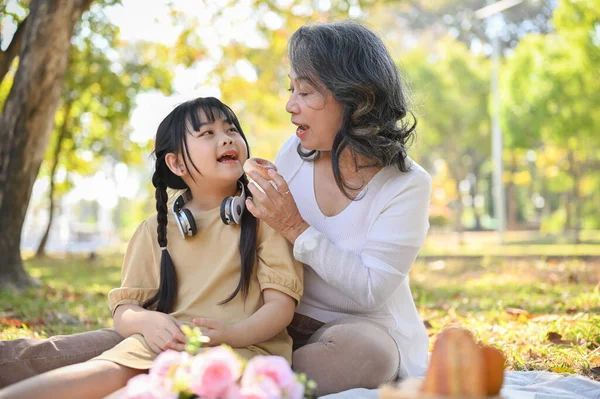 Happy and caring Asian grandmother feeding doughnut to her little granddaughter while picnicking in the beautiful park together.