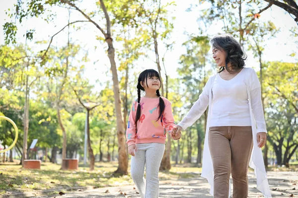 Happy and caring Asian grandmother holding her granddaughter\'s hand while walking in the beautiful summer park together. family bonding, happiness, outdoor activity