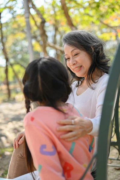 Caring and kind Asian grandmother tells a story to her granddaughter while relax on a bench in the park together.