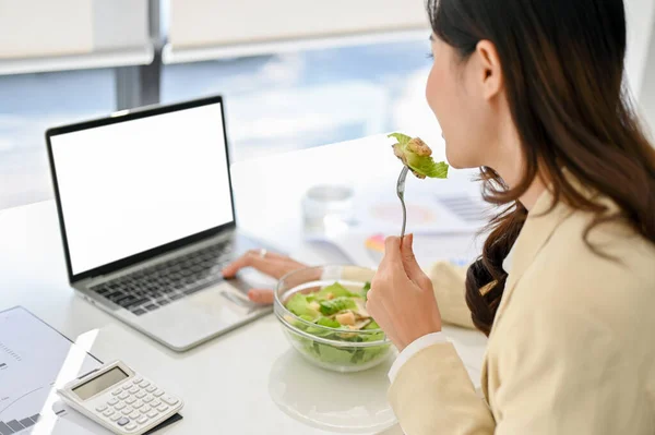close-up image of a busy millennial Asian businesswoman eating healthy salad bowl, having lunch while working on her project on laptop in the office. laptop white screen mockup, hardworking, multitask