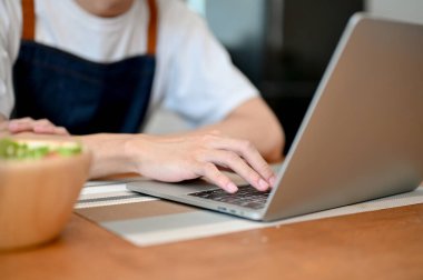 close-up image of a man in a apron young laptop computer at dining table in the kitchen, searching food recipe.