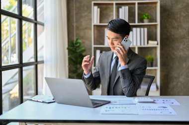 Professional and smart millennial Asian businessman or male business consultant is on the phone with his client, counseling business over the call, working in his office.