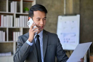 Handsome and professional millennial Asian businessman in formal business suit looking at the document while on the phone with his client.