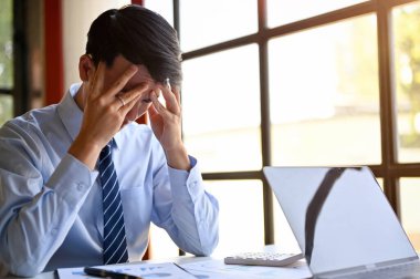 Stressed millennial Asian businessman sits at his desk, worried about his business's financial investment outcome, getting trouble from clients, or dissatisfied with work's performance.