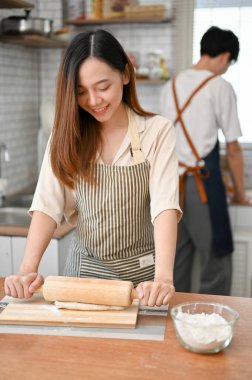 Happy young Asian female in apron is kneading raw dough with a rolling pin, making dough while her boyfriend is preparing ingredients in the kitchen.