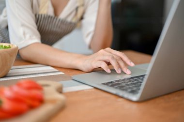 Close-up hand image of a female in apron using laptop computer, typing on keyboard, searching food recipe on internet at her kitchen table.
