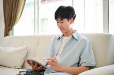 Side view of a happy young Asian man in casual clothes relaxing on sofa in his living room and using his digital tablet.