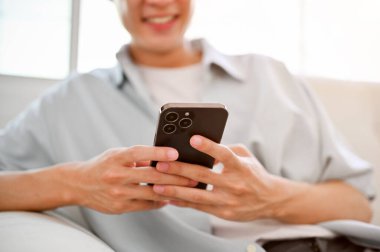 close-up image of a happy young Asian man relaxing on sofa in his living room, using his phone, texting with his girlfriend.