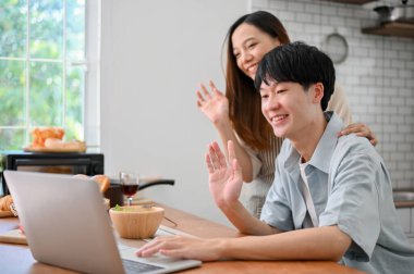 Side view image of happy young Asian couple waving hands, saying hi to their parents through a video call on laptop in the kitchen.