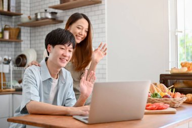 Happy young Asian couple waving hands, saying hi to their parents through a video call on laptop in the kitchen, spending free weekend together at home.