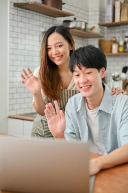 Portrait, Happy and smiling young Asian couple waving hands, saying hi to their parents through a video call on laptop in the kitchen.