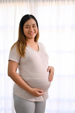 Portrait of a happy and beautiful young Asian pregnant woman in casual comfy clothes stands by the window, touching her belly with love.