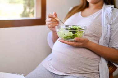 Close-up image of an Asian pregnant woman in comfy clothes eating a healthy salad for breakfast at a table by the window. pregnancy, motherhood lifestyle
