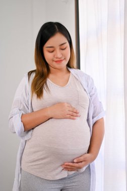 Relaxed and happy Asian pregnant woman in casual clothes feeling her baby, touching her belly with love and care. parenting and motherhood concept