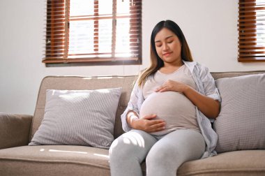 Happy Asian pregnant woman in comfortable clothes relaxes on the sofa in her living room, touching and feeling her baby in her belly with love. motherhood concept