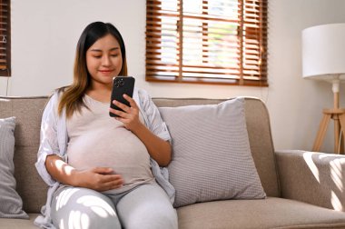 Happy Asian pregnant woman using her phone, reading an online column about motherhood preparation while relaxing on sofa in her living room. motherhood, pregnancy lifestyle 