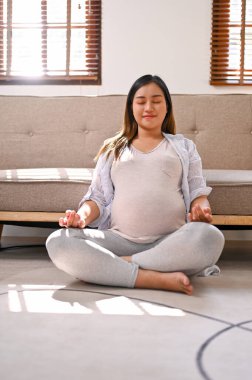 Portrait of a happy and relaxed Asian pregnant woman in comfortable clothes meditating in her living room.