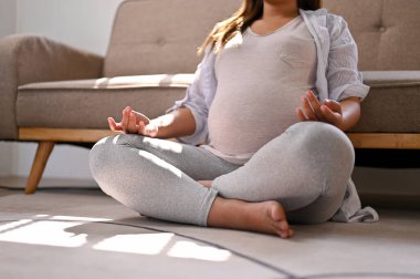 Cropped image of a happy and relaxed Asian pregnant woman in comfortable clothes meditating in her living room, lotus pose, concentrating breath.