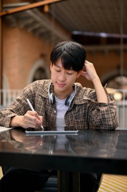 Portrait of a focused young Asian man in casual clothes designing his artwork or taking notes on a digital tablet, remote working at the cafe.