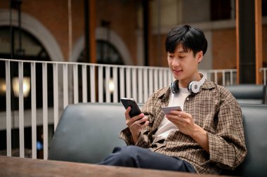 Happy Asian man in casual clothes holding a smartphone and a credit card, using mobile banking application, registering credit card payment on shopping app.