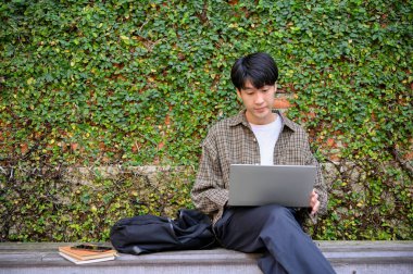 Smart and focused young Asian male college student or freelancer remote working in the city park or garden, using his portable laptop on a bench, working on his tasks.