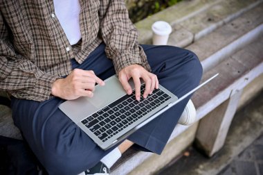 Top view of an Asian man in casual clothes sitting on bench in the park and using his portable laptop, remote working outdoor.