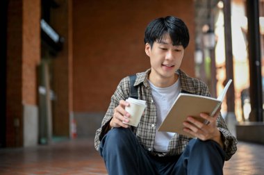 Handsome and smart young Asian male college student in casual clothes sitting on stairs outside of the campus building, holding a coffee cup and reading a book.
