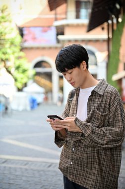 Portrait of a handsome Asian man in casual clothes using his smartphone, texting to someone or using mobile application while waiting his friends at the city street.
