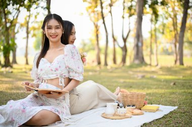 Çekici ve çekici, güzel elbiseli Asyalı kadın arkadaşıyla piknik elbisesinde oturup yeşil parkta piknik yapmaktan hoşlanıyor.