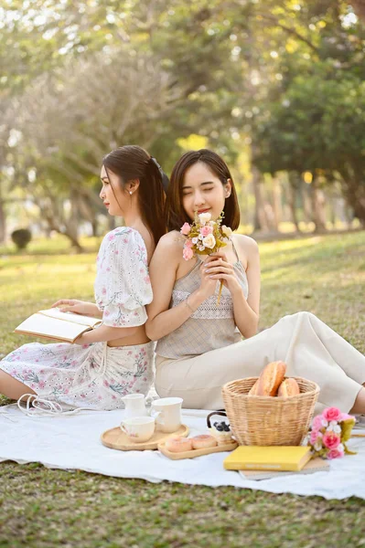 Charming beautiful young Asian woman in a lovely dress sits on a blanket, holding a flower bouquet, enjoying a picnic with her friend in the park.
