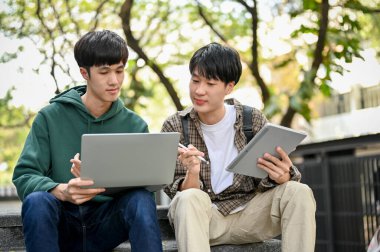 Two smart young Asian male college students focusing on his school project, looking at laptop screen, using laptop and tablet, discussing and working together at the campus park.
