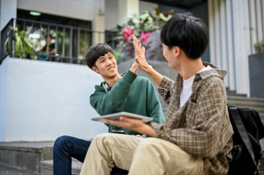 Cheerful young Asian male college student giving high five to his friend, sitting at the outside of the campus building together. greeting gesture