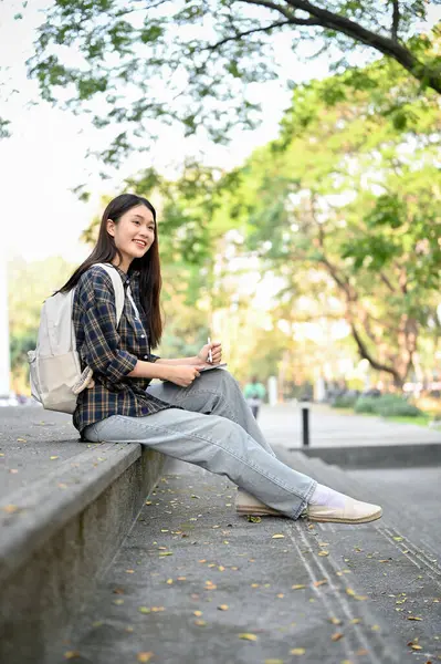 Charming young Asian female college student in trendy clothes with her portable tablet sits on street stairs, relaxing after classes.