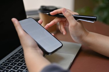 Close-up image a businesswoman holding pen and using her smartphone while working on her business tasks on laptop computer.
