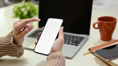 A businesswoman working from home, using her smartphone at her working desk. smartphone white screen mockup. close-up image