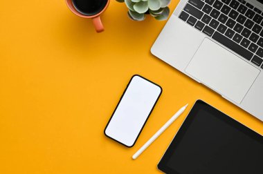 Top view of a creative office desk workspace with smartphone white screen mockup, stylus pen, tablet, laptop, coffee cup and decor plant on yellow background.