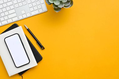 Top view of a modern office desk in yellow background with smartphone white screen mockup, pen, book, keyboard computer, decor plant and copy space.