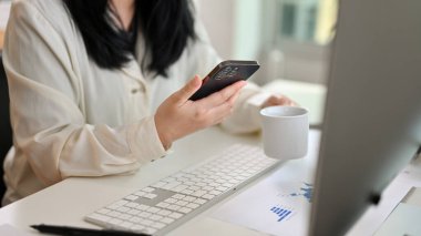 An Asian businesswoman or female manager sipping coffee and using her smartphone at her desk in the office. cropped image