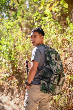 Portrait of an active Asian male traveler with backpack and trekking gear trekking in the forest on the sunny day alone, exploring in the forest. adventure outdoor activity concept