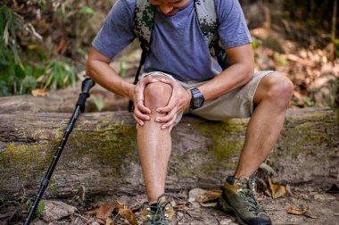 Cropped image of a stressed Asian male trekker sits on a wooden log and holds his knee, suffering from knee pain, severe leg pain, injured during trail. accident, health problems concept