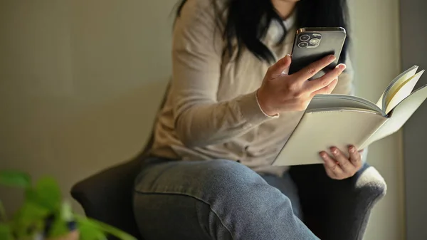 A young Asian woman in casual clothes using her smartphone and reading a book while relaxing in the cafe. cropped image