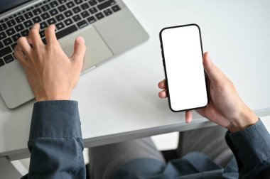 Top view of a businessman working at his office desk, using laptop computer and holding a smartphone. smartphone white screen mockup